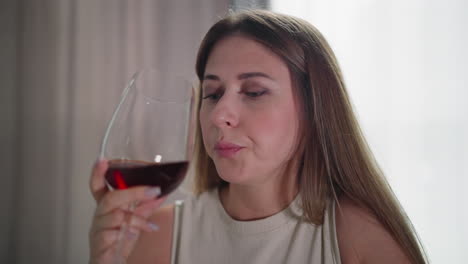 young woman in sleeveless outfit drinks red wine from glass while gazing thoughtfully at cup, soft lighting highlights serene indoor setting with neutral curtains in background