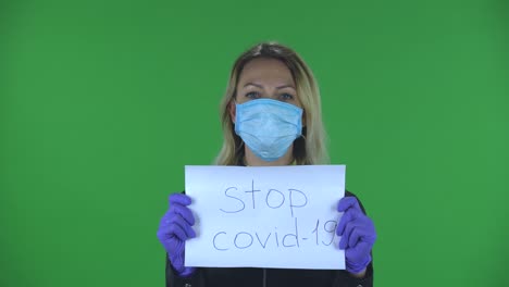 portrait of beautiful young woman in medical mask is looking at camera and holding a poster stop kovid-19.blonde with loose hair in a black jacket and jeans on a green screen in the studio. health protection corona virus concept