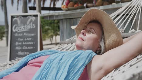 Relaxed-senior-caucasian-woman-lying-on-hammock-sleeping-in-the-sun-by-beach-bar,-in-slow-motion