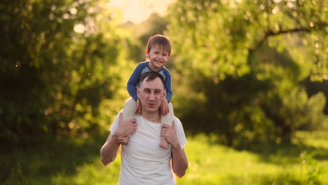 a child sitting on the neck of his father while walking in summer field at sunset.