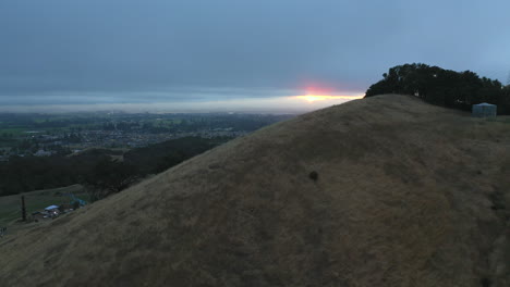 Aerial-flyover-of-a-grassy-ridge-with-the-sunset-just-peaking-below-the-clouds-with-the-east-part-of-Santa-Rosa,-California-in-the-background