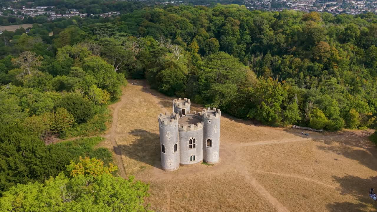 Cinematic drone orbit of historic Blaise Castle in Henbury, Bristol. Elliptical movement reveals the Gothic folly tower surrounded by woodland clearing with visible pathways through dry grass