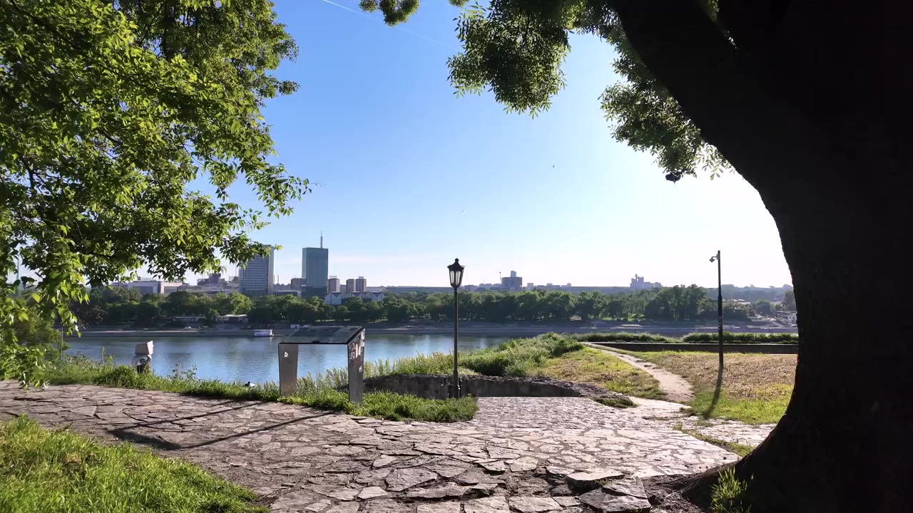 Un paseo por un parque junto al río, rodeado de árboles. El sol brilla en el cielo despejado mientras la hierba verde crece junto al sendero. La gente disfruta del aire libre a lo largo de la orilla del río.