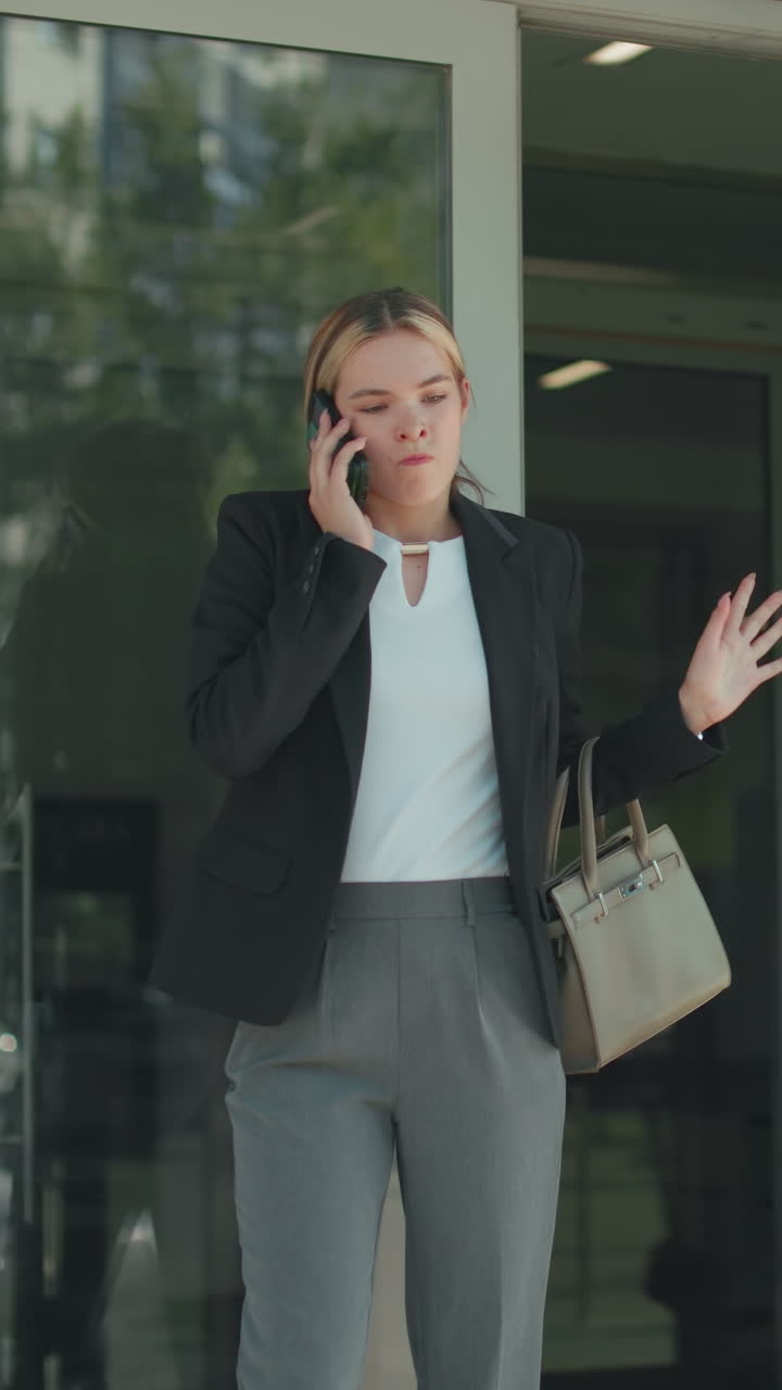 Furious lady on phone drops device in frustration while continuing call, exiting building in professional outfit, holding handbag, urban glass structure reflects cars and cityscape