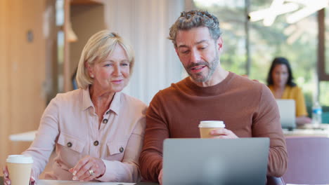 mature female businesswoman brings takeaway coffee for male colleague as they have meeting with laptop