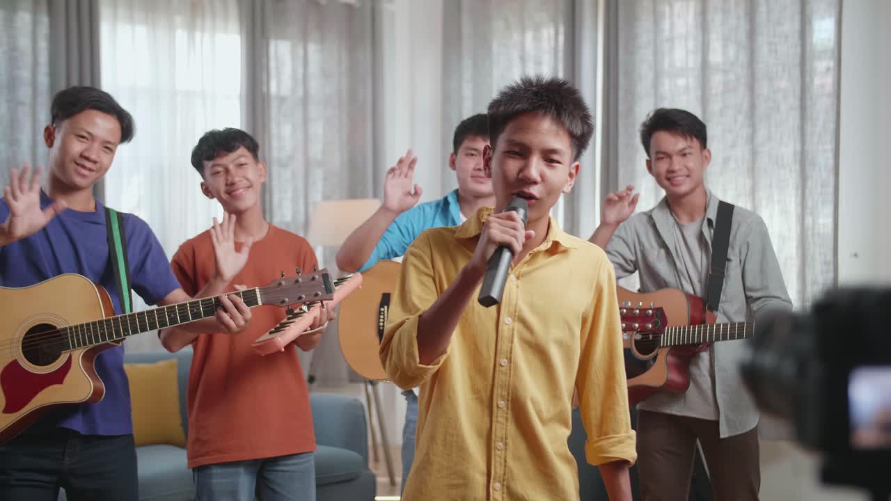젊은 아시아 친구 그룹이 악기를 들고 집에서 라이브 스트리밍을 하는 동안 손을 흔들고 카메라에 말하고 있습니다.