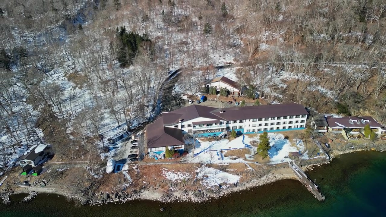 Luftbåren panoramisk sporingsvisning af et hotelkompleks ved søen, beliggende blandt bladløse vintertræer og sne i Hokkaido.