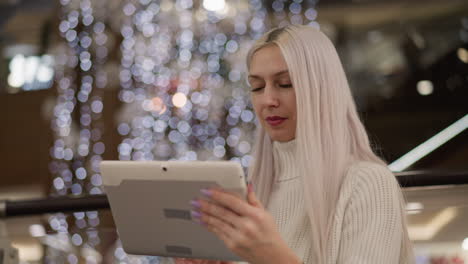 shoulder view of mall goer seated on bench near glass railing under ambient bokeh lights, her focus on touchscreen tablet as she taps and smiles, cozy sweater accents casual tech lifestyle moment