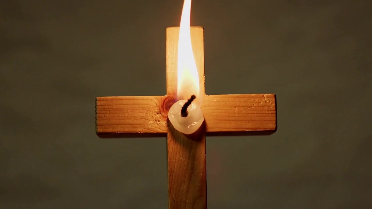 Zooming camera showing white candle on wooden cross burning, melting wax in quiet room for closeup