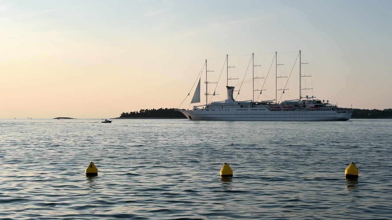 A luxury cruise ship sails past Rovinj, Croatia during a serene sunset. Calm Adriatic waters reflect soft golden light and silhouettes of the coastline