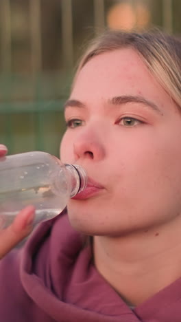 joven con capucha marrón bebiendo agua de una botella después del entrenamiento, capturada en perfil lateral, con la valla de la barra borrosa en el fondo, momento refrescante después del ejercicio