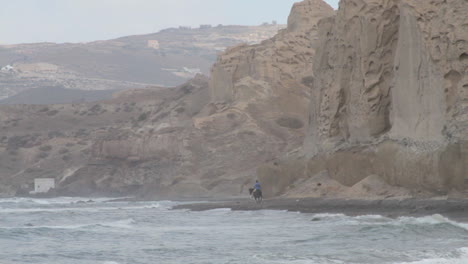 Teleaufnahme-Eines-Reiters,-Der-Auf-Seinem-Schwarzen-Pferd-Am-Schwarzen-Strand-Von-Santorini-Reitet