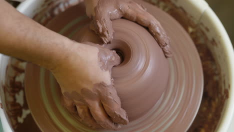 Close-up-skilled-potter-fingers-forming-wet-clay-shape-spinning-on-workshop-pottery-wheel