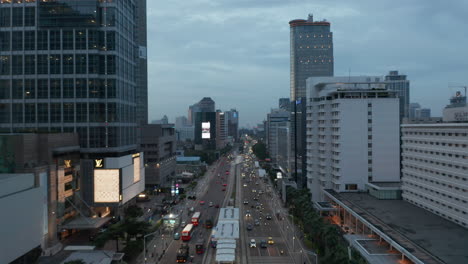 Toma-Aérea-De-Carro-De-Bajo-Vuelo-Del-Tráfico-De-La-Ciudad-En-El-Moderno-Centro-De-La-Ciudad-Al-Atardecer-En-Yakarta,-Indonesia