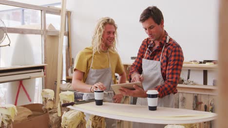 Two-Caucasian-male-surfboard-makers-standing-and-working-on-projects-using-a-tablet