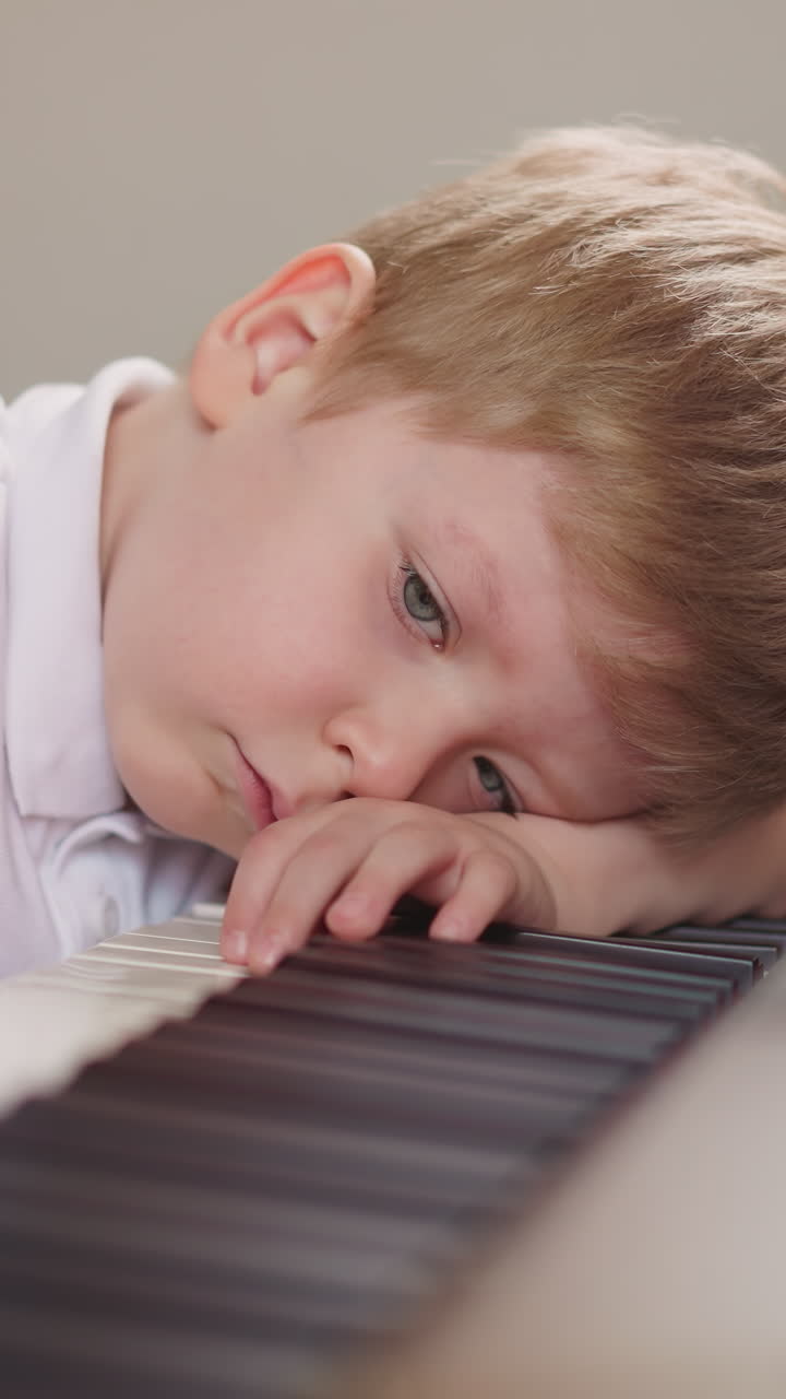 Fatigued toddler boy puts head on keyboard and presses random keys on electric piano in class closeup. Little child disliking classic music suffers during lesson