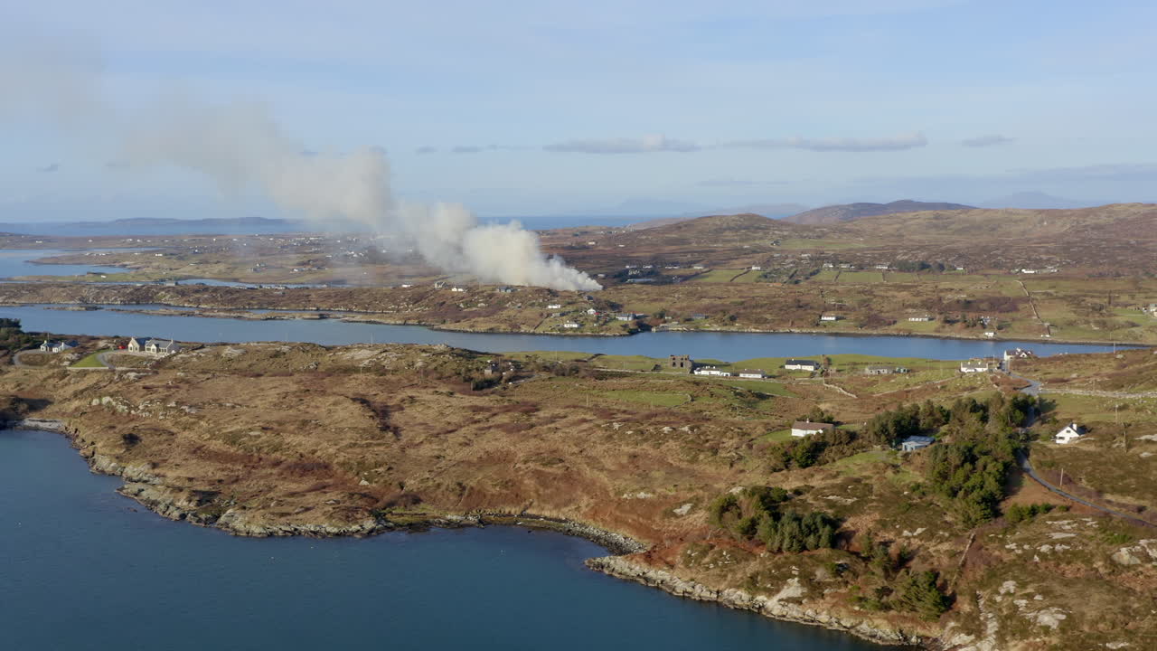 Wide aerial pan over Claddaghduff, with smoke rising from farmland in the distance. Connemara