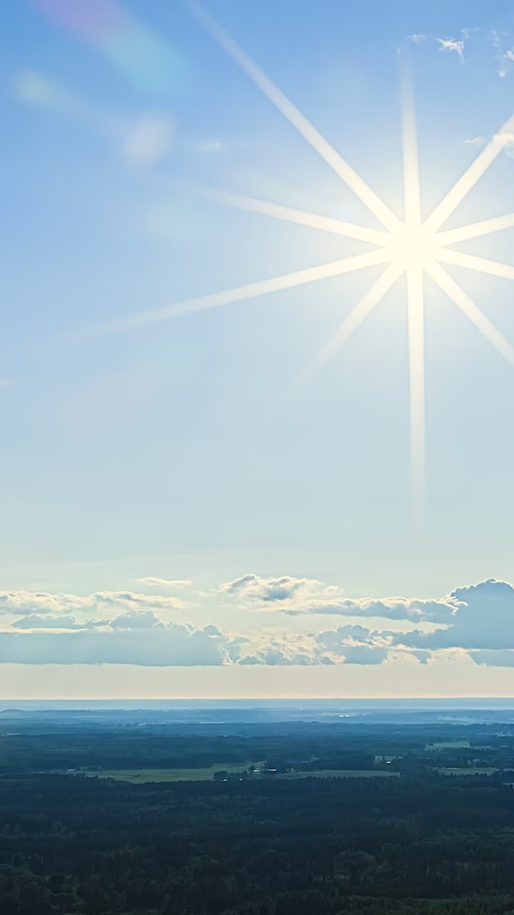 地平線に沿って広がる遠方の田園地帯の農地と森林を、明るい太陽の光線が照らす。空撮の縦長構図。
