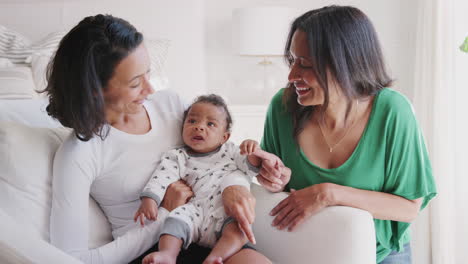 Millennial-mother-sitting-in-armchair-holding-her-baby-son,-her-mother-beside-them