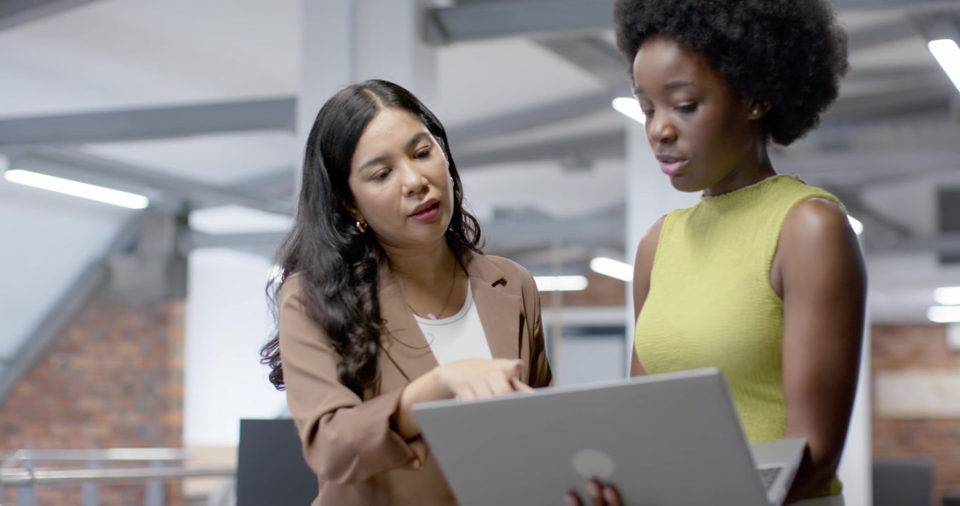 Diverse women coworkers working in office, right showing laptop left with blue mug tapping trackpad