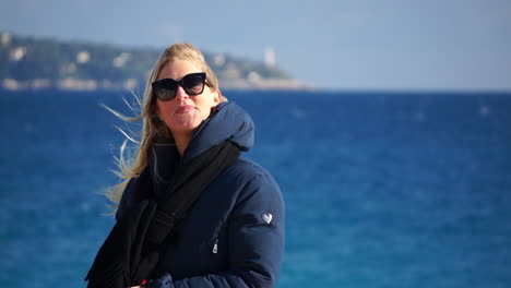 a young blonde woman laughs to the camera with the blue sea in soft focus behind her on a windy, sunny day slomo