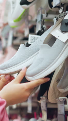 dama de vestido rosa en la tienda de zapatos examina un par de zapatillas de ceniza de cerca, centrándose en la suela mientras que varios otros zapatos se muestran en el estante ligeramente borroso, la tienda está bien iluminada