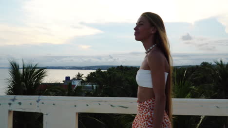 young girl in white crop top walks on a balcony with beach at far end at sunset