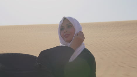 muslim woman in white hijab and traditional black dress sitting on sand in a windy desert and smiling at camera