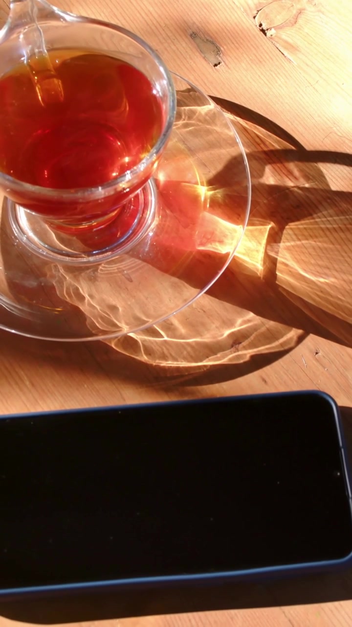 Video verticale: Telecamera che si inclina rivelando una tazza di vetro su un piattino su un tavolo, con caustiche del sole in movimento e un telefono.