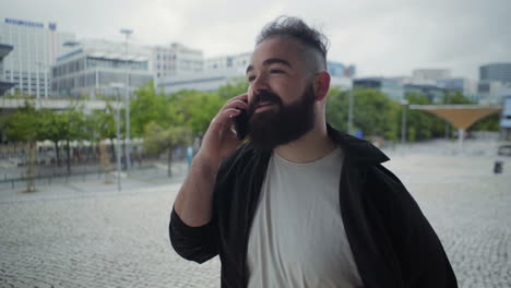 hombre barbudo sonriente paseando por la calle mientras habla por teléfono.