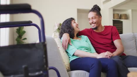feliz pareja biracial sentada en el sofá abrazándose en la sala de estar, con silla de ruedas en primer plano