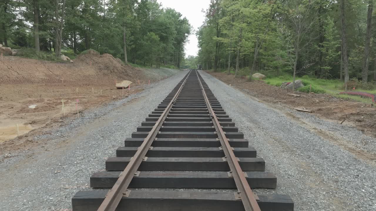 Workers lay new train tracks in a forest area. The ground is prepared with gravel and wooden beams. Train Slowly Travels in Trees line the sides of the track. Work is in progress during the day