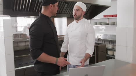 cocinero caucásico enfocado instruyendo a un cocinero aprendiz usando una computadora portátil en la cocina, en cámara lenta