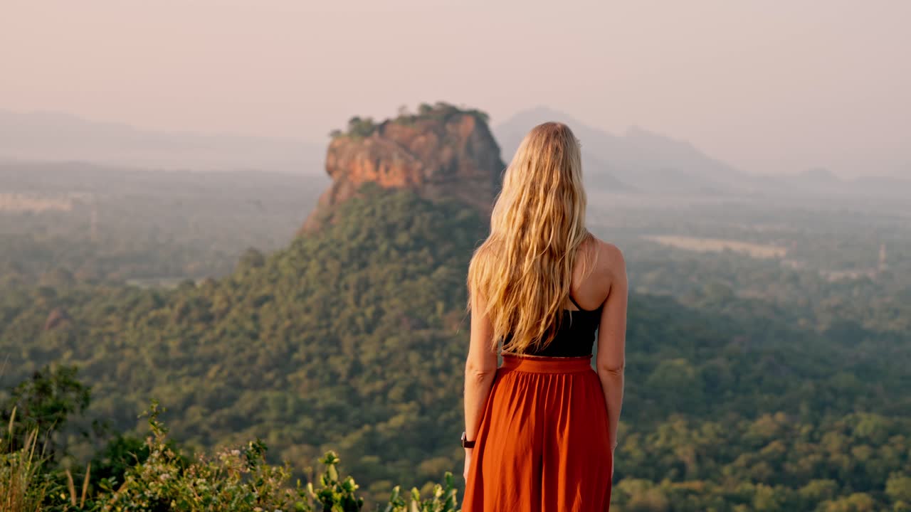 A woman walks peacefully with the majestic Sigiriya Rock landmark visible in the background during a vibrant sunrise in Sri Lanka.