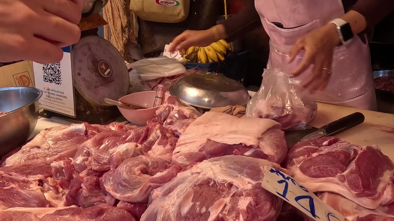 A butcher packages fresh cuts of meat at a market stall, using a plastic bag and scale.
