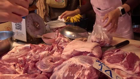 a butcher packages fresh cuts of meat at a market stall, using a plastic bag and scale.