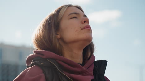 strong willed trainee in hoodie with eyes closed stretches neck round with relief while warming up outdoors against urban sunlit background with hair illuminated and serene focus before workout