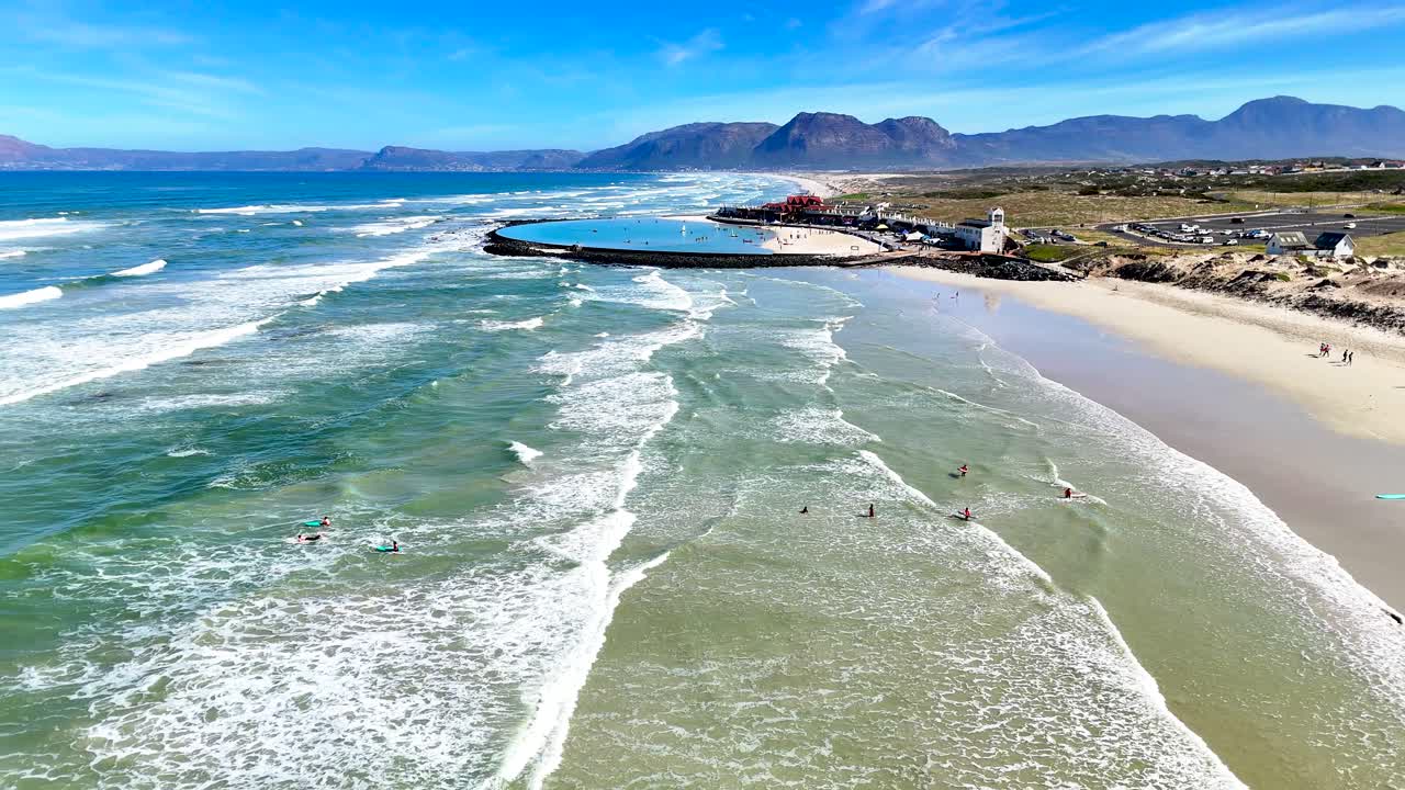 Drone clip of a stretch of beach with a tidal pool in the background and swimmers enjoying the waves of the ocean in Cape Town South Africa