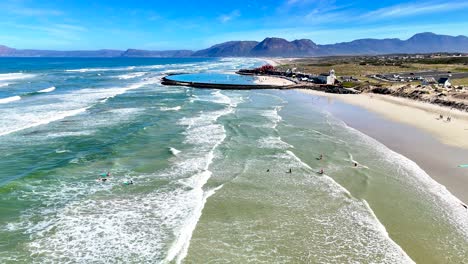 drone clip of a stretch of beach with a tidal pool in the background and swimmers enjoying the waves of the ocean in cape town south africa