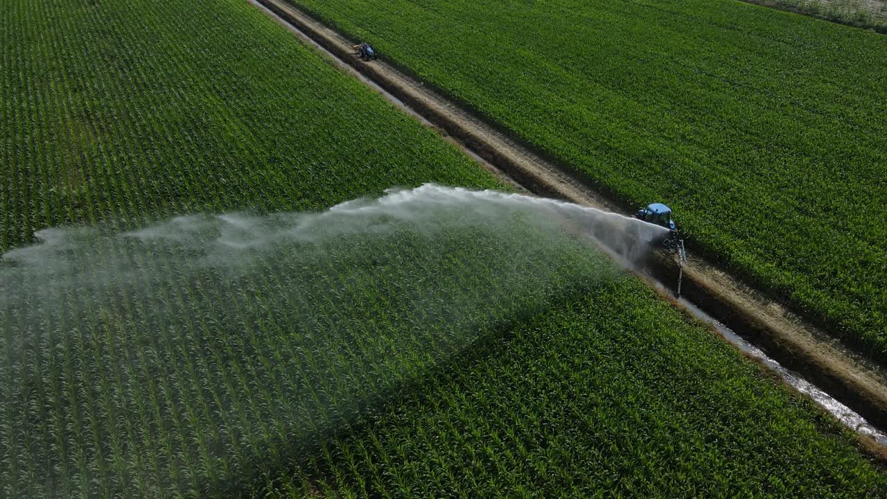 Dynamic 4K drone aerial rotating clockwise above a tractor irrigating farmland in Italy, with a tilted gimbal showing the crop and water spray from a near top-down perspective