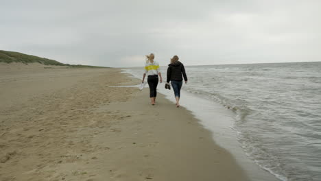 Rear-shot-of-two-female-girlfriends-walking-at-seashore-during-holiday-trip-in-Netherlands