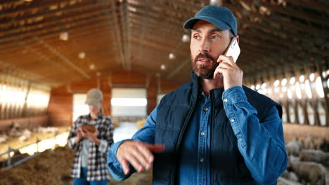 close-up view of young caucasian man in cap walking in stable with sheep flock and talking on mobile phone