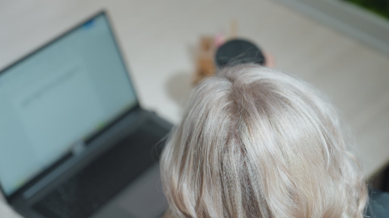 Top down view of woman working on laptop pauses typing to sip from black cup then resumes, seated at desk with visible screen, organized workspace, blonde hair lit by indoor light