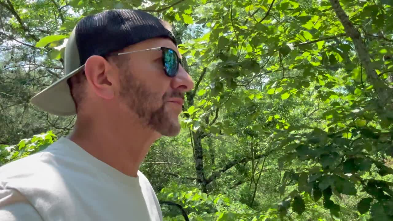 Bearded man wearing sunglasses and a backwards baseball cap on a nature hike in a forest during summer - slow motion portrait
