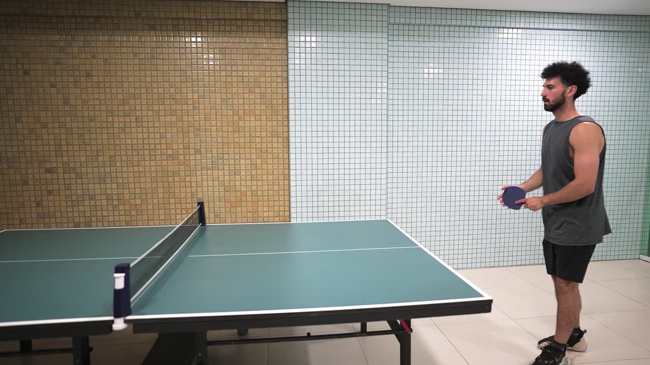 A left-handed player serves in an indoor table tennis match, captured in a static wide-angle shot. The frame highlights his athletic stance, controlled movement, and energetic lifestyle