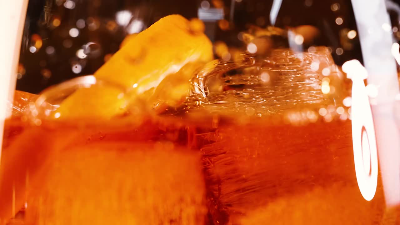 Vista macro de líquido laranja enchendo um copo com cubos de gelo e guarnição cítrica.