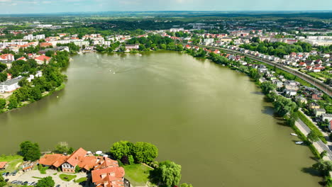 Aerial-view-of-Iława,-showcasing-a-large-lake-surrounded-by-residential-and-historic-buildings,-lush-greenery,-and-a-tranquil-summer-atmosphere