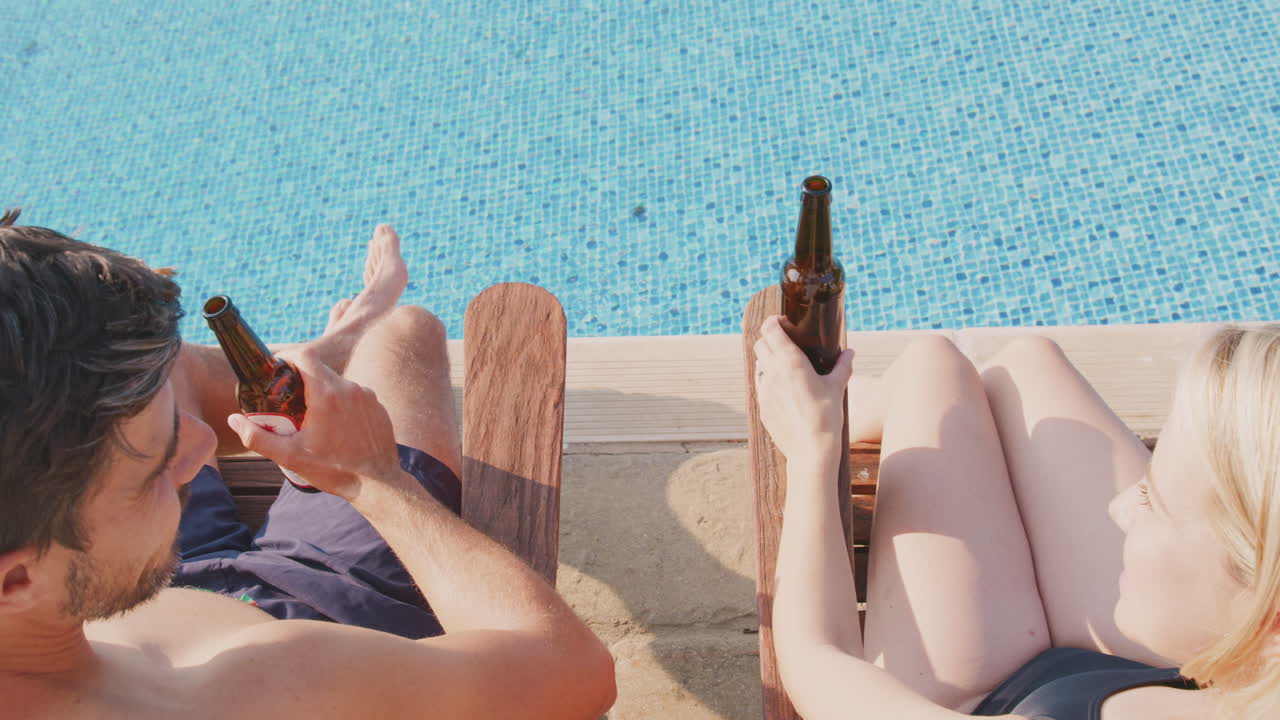 Rear View Of Couple On Summer Vacation Sunbathing On Loungers And Drinking Beer By Swimming Pool