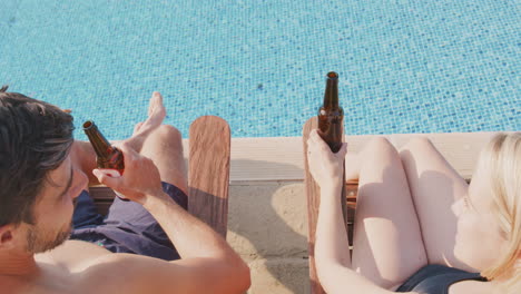 Vista-Trasera-De-Una-Pareja-En-Vacaciones-De-Verano-Tomando-El-Sol-En-Las-Tumbonas-Y-Bebiendo-Cerveza-Junto-A-La-Piscina.