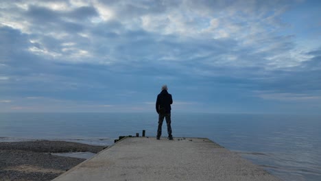 Nahaufnahme-Eines-Mannes,-Der-Am-Ende-Eines-Stegs-In-Der-Abenddämmerung-In-Zeitlupe-Am-Fleetwood-Beach-In-Lancashire,-Großbritannien,-Auf-Das-Meer-Starrt